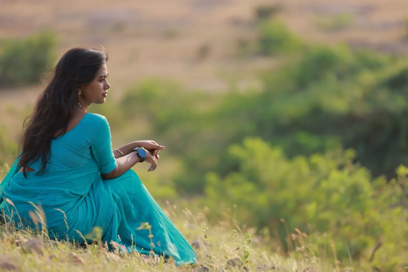 A woman in a teal saree sitting on a grassy hillside, looking out calmly.